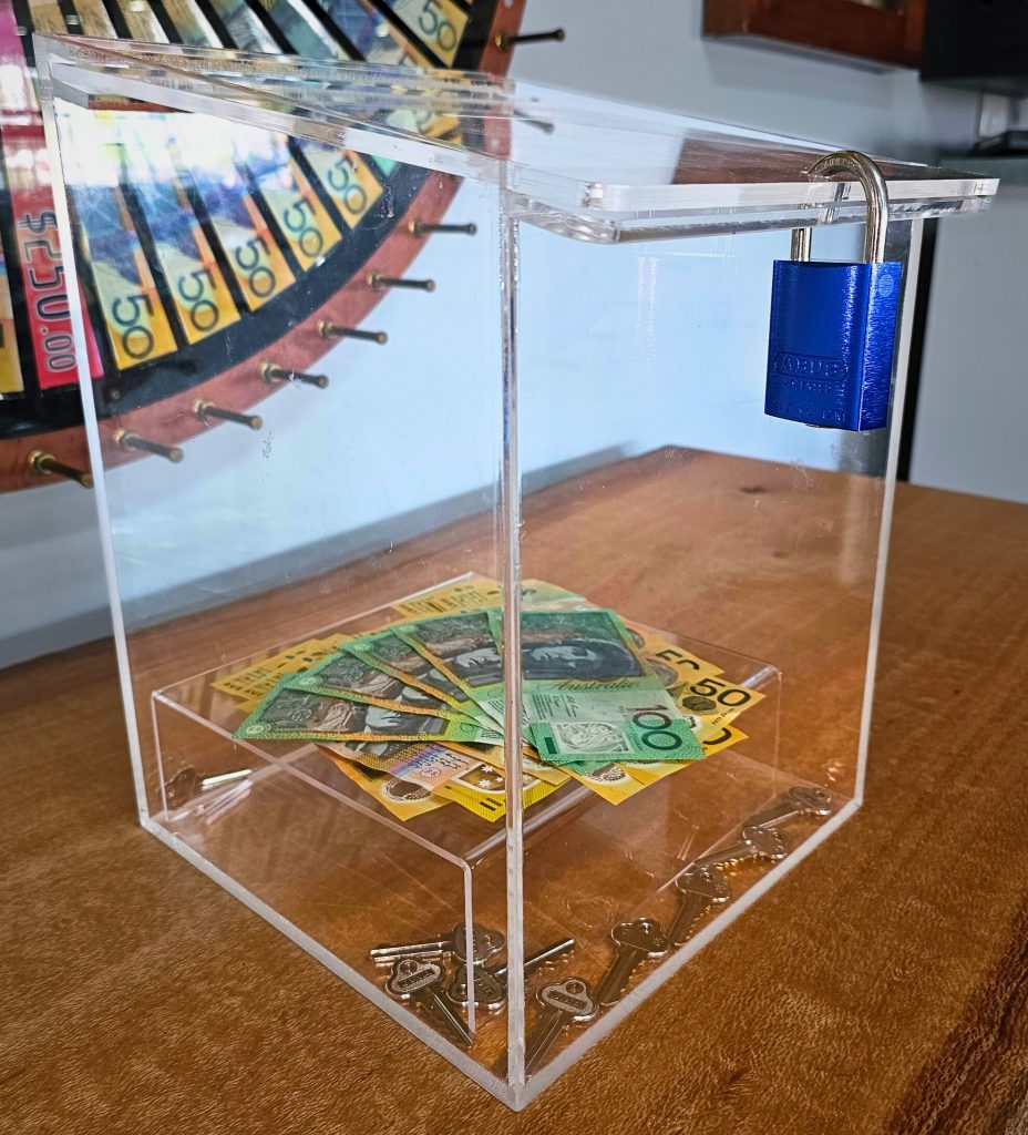 A clear acrylic box with a blue lock, holding fanned $100 and $50 notes on top of a pile of keys